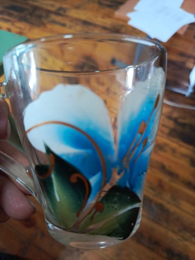 A close-up, eye-level shot shows a clear glass mug with a painted blue flower design and green leaves. The mug is held by a person's hand, with the thumb and fingers visible on the left side of the image. The mug is positioned on a wooden surface, likely a table, with a textured grain. In the background, out of focus, are some white papers and a blurred green object. The lighting suggests an indoor setting. The location is not specified in the image or provided context.