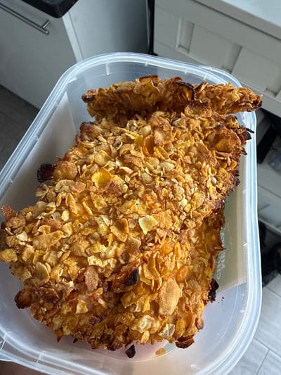 The image shows a close-up view of a piece of fried chicken coated in cornflakes. The chicken is a golden brown color, with some pieces appearing slightly darker and more charred. It is resting inside a clear plastic food container. The container has a white lid that is partially visible at the top of the image. The background is out of focus, but appears to be a kitchen or dining area with white cabinets. There is a label on the inside of the container lid, but the text is mostly obscured and unreadable, though the word "CHOCE" is partially visible. The lighting suggests it is daytime, likely indoors. The scene is static, with no people or activity visible.