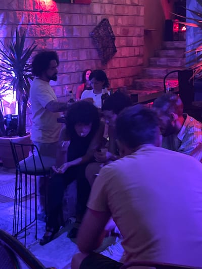 The image shows a group of people gathered at a venue with dramatic purple and pink lighting. The setting appears to be an outdoor or semi-outdoor space with stone walls and stairs in the background. A large potted plant with spiky leaves is visible on the left side of the frame.
In the foreground, a person wearing a light-colored t-shirt is seated, with their back to the camera. To their left, another person with dark, curly hair is seated and appears to be smoking a cigarette. To the left of them, a man with dark, curly hair and a beard stands and looks towards the seated individuals. In the background, partially obscured by the foreground subjects, are two other individuals, one appearing to be a woman in a white top and another person whose face is not clearly visible.
The overall atmosphere is dimly lit and social, suggesting a bar, restaurant, or party setting. The location is identified as Herceg Novi, Montenegro, implying a scene typical of its vibrant nightlife or social gatherings, particularly during evening hours. The lighting and the casual arrangement of people suggest a relaxed, informal gathering.