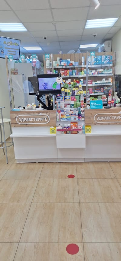 The image depicts the interior of a pharmacy in Nakhodka, Russia. The scene is dominated by a counter with shelves filled with various medicines and health products. A computer monitor is visible on the counter, displaying an animation of two mice. A transparent protective screen is installed on the counter, likely for hygiene purposes. On the counter, the word "ЗДРАВСТВУЙТЕ" (hello in Russian) is prominently displayed on a wooden panel. There are also yellow circular stickers on the floor, indicating social distancing markers. The floor is tiled with a light brown wood-like pattern. The overall environment appears to be clean and organized.