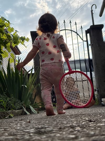 In Oreshak, Bulgaria, a young child stands on a paved walkway, facing away from the camera, holding a child-sized pink badminton racket. The child wears a white t-shirt adorned with strawberries and pink patterned leggings. To the left, lush green plants and a fence with a house in the background are visible. Behind the child, a black metal gate stands open, with a car partially visible through it. Another young child is lying on the ground behind the gate, their legs and torso visible through the racket's netting. The scene is illuminated by natural light, suggesting it is daytime, with clouds present in the sky.