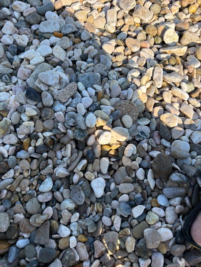 This media file is a close-up, overhead shot of a beach covered in various sizes and colors of smooth pebbles. The pebbles range from white, beige, and light gray to darker grays, browns, and even some with speckled patterns. Sunlight casts distinct shadows across the stones, indicating a sunny day. In the upper right corner, a small portion of a person's foot, wearing a dark sandal, is visible, suggesting the photo was taken by someone standing on or near the beach. The scene is a natural outdoor setting, likely a beach or shoreline. The texture of the pebbles is varied, with some appearing almost polished and others rougher. The lighting creates a strong contrast between the illuminated and shadowed areas, highlighting the three-dimensional nature of the pebble-covered surface. The location is identified as Općina Baška, Croatia. No text is visible in the image.