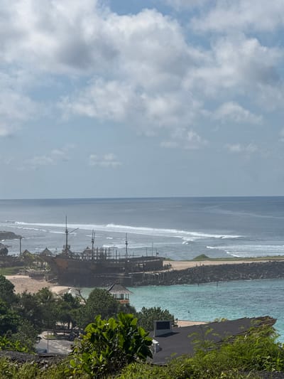 An eye-level, outdoor shot looking out to sea in Ungasan, Indonesia. The sky is overcast. The ocean has rough waves. A structure of wooden poles is present in the distance, near the shore. Beach foliage and trees are in the foreground. A concrete parking lot is visible.