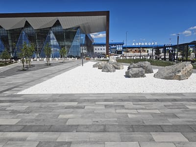The image shows the Blagoveshchensk Airport, located in Blagoveshchensk, Russia. The airport's modern, angular architecture is visible, with a large glass facade reflecting the blue sky and clouds. In the foreground, a pathway made of gray paving stones leads to a white gravel area featuring several large rocks. Beyond the gravel, a row of young trees stands in front of the airport building. In the background, the airport terminal building displays the word "АЭРОПОРТ" (Airport) in large white letters against a blue backdrop. Several vehicles, including cars and a bus, are parked near the terminal. The scene is set under a clear blue sky on a sunny day.