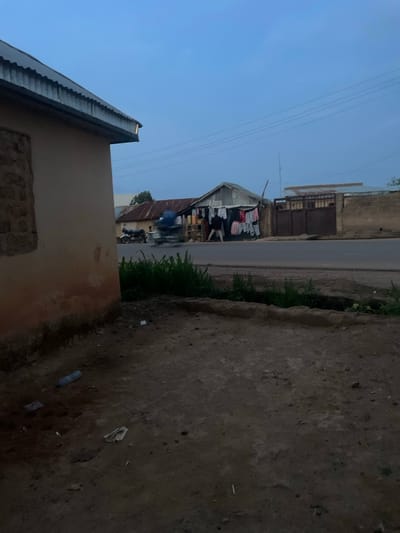 The image shows a street scene in Reifild-Ist, Nigeria. A road runs horizontally across the middle, flanked by buildings on either side. On the left, clothes are hanging outside of a building to dry. A person on a motorcycle is on the road in the direction of the clothing. A building is in the foreground on the left side of the image. In the foreground of the right side is what appears to be a sidewalk that leads to a grass patch beside the road. The sky is visible above the buildings.
