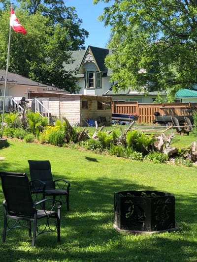 The image depicts a backyard scene in the city of Severn, Canada, on a sunny day. In the foreground, two dark-colored patio chairs are positioned on a well-maintained green lawn, with a black metal fire pit situated between them and slightly further back. The fire pit has intricate cut-out designs.

Behind the chairs, a landscaped garden area features a variety of plants, including ferns, tall grasses, and yellow flowers, interspersed with weathered driftwood. A small, light-colored wooden shed is visible in the middle ground, with a red-trimmed roof and two small windows. To the left of the shed, a white deck with railings and stairs leads up to a house. A Canadian flag flies proudly from a flagpole on the left side of the property.

Further back, a wooden fence encloses the yard. Several boats are visible parked behind the fence, including a blue aluminum boat on a trailer and a darker aluminum boat with an outboard motor. In the background, several houses with green roofs are partially obscured by mature, lush green trees. The sky is clear and blue.