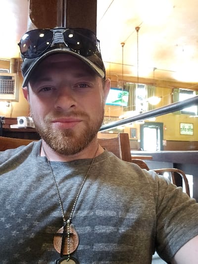The image depicts a single male subject seated indoors, facing the camera. The exact location within Centralia, United States, is not discernible from the provided visual information.
**Subject:**
A Caucasian male, appearing to be in his 20s or 30s, occupies the central foreground. He has a reddish-brown beard and is wearing a dark-colored baseball cap with dark sunglasses resting on top of the brim. His eyes are open, and he is looking directly forward. He wears a grey, short-sleeved t-shirt with a faded, horizontally striped pattern that resembles the American flag. A silver-colored bead chain necklace hangs around his neck, supporting multiple pendants: a dark cross, a circular copper-toned medallion with an etched design, and a smaller, dark circular object suspended beneath the copper piece.
**Environment and Objects:**
The subject is seated in what appears to be a casual public establishment, possibly a restaurant or bar, indicated by the interior decor. A dark wooden chair back is visible behind his right shoulder. The background features light-colored walls, possibly painted yellow or tan, with dark wooden structural elements. The ceiling is light-colored, featuring multiple pendant light fixtures.
To the left of the subject, a white rectangular air conditioning unit or vent is mounted on a wall, with a smaller white electronic device positioned below it on a counter. In the mid-ground, a dark horizontal bar or railing runs across the frame. Further into the background, a television screen displaying a bright blue sky and green landscape is visible. Windows with what appear to be green curtains or blinds are also present. A dark doorway is discernible on the right side of the background. The floor is not visible. Lighting appears to be a mix of natural light from windows and artificial overhead lighting.