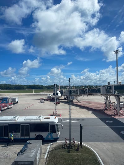 This image captures an airport scene in Mai Khao, Thailand, under a partly cloudy blue sky. In the foreground, a white bus with blue accents, labeled "AOTGA PB-042", is parked. A person wearing a yellow vest and blue shirt is walking near the bus. To the left of the bus, airport luggage carts are visible, some with red markings and identification numbers. A jet bridge extends from the right side of the frame to an unseen aircraft. The tarmac is marked with red and white barriers and lines. In the middle ground, an airplane is parked, connected to the jet bridge. Several service vehicles, including a small white truck, are on the tarmac. Beyond the tarmac, a green grassy area transitions into a dense line of trees and foliage, with hills visible in the distance. A tall light pole stands on the far right. The overall environment appears to be a sunny day at an international airport.
