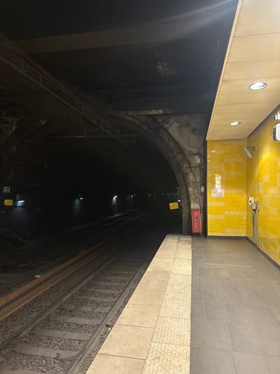 This image depicts an underground train station platform and associated tunnel, located in Vincennes, France. The scene is devoid of human presence or activity.
On the left side of the frame, two parallel railway tracks, supported by ballast, recede into a dark, concrete-arched tunnel. The tunnel interior is dimly lit by scattered overhead lights, revealing multiple sets of cables and conduits running along the arch structure and ceiling. Several rectangular yellow warning signs with dark text/symbols are affixed to the tunnel wall at intervals.
The platform, occupying the right side of the image, is brightly illuminated by multiple recessed circular lights embedded in a flat, light-colored ceiling panel. Its floor is paved with large grey tiles, featuring a distinct light-colored tactile strip with raised circular bumps running along the edge adjacent to the tracks. The platform wall is faced with horizontally-arranged bright yellow ceramic tiles, displaying subtle variations in shade between horizontal bands. Essential objects mounted on this wall include a prominent red fire extinguisher contained within a red casing, positioned near the junction of the tunnel arch and platform wall. Further along the yellow tiling, a grey emergency communication box with a handset is mounted, alongside two adjustable wall-mounted light fixtures with white casings, directed towards the platform. A white rectangular information panel or map is also present on the yellow tiled wall above the fire extinguisher.