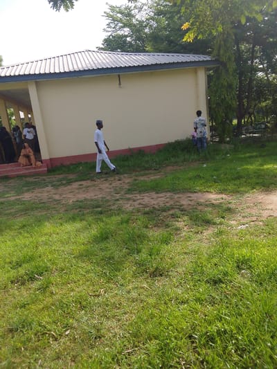 The image depicts an outdoor scene in Bauchi, Nigeria, featuring a small, light yellow building with a grey, corrugated metal roof. A covered walkway extends from the left side of the building, with several individuals gathered within its shaded area. On the right side of the building, a person wearing a white traditional outfit and a cap walks away from the camera. Further to the right, another individual in a patterned shirt and blue jeans stands near a tall, leafy tree. The ground in front of the building is a mixture of dirt and patchy green grass. In the background, more trees and a park bench are visible, suggesting a public or communal space. The lighting indicates it is daytime, with shadows cast by the building and trees.