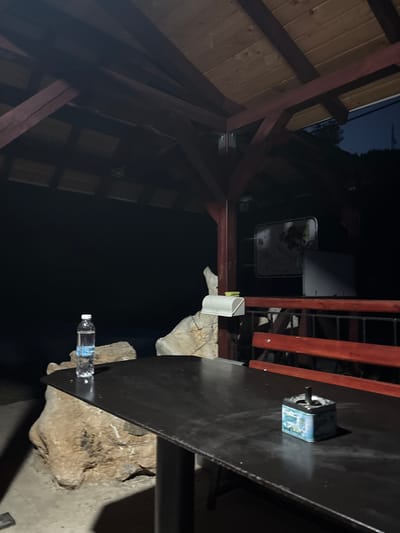 A dark, outdoor scene is depicted, likely at night, under a wooden gazebo with exposed beams. The foreground is dominated by a dark, rectangular table. On the table, towards the right, sits a small, square, light blue metal ashtray with an image of a van on it and a black object, possibly a cigarette butt, protruding from it. To the left on the table, a clear plastic bottle of water with a white cap and a blue label is standing upright. The table is supported by dark, slender legs, and its surface appears slightly reflective.

Behind the table, a large, irregularly shaped rock formation is visible, partially supporting the table's edge. Further back, a red wooden bench is positioned behind a dark railing, which is also made of wood and metal. Above the bench, a rectangular, framed object with an image on it hangs on what appears to be a wall or partition, illuminated by a faint light. The background is very dark, suggesting it is nighttime, with the faint outline of trees and possibly a structure visible in the distance against a dark sky. The overall atmosphere is still and quiet. The location is identified as Pechinska, Bulgaria.
