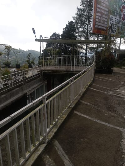 The scene depicts an outdoor, multi-level terraced area under an overcast sky, with the stated location being Siruppiddy, Sri Lanka. However, a large commercial billboard prominent on the right displays text in Indonesian, including "DIJUAL" (FOR SALE), "DISEWAKAN" (FOR RENT), and the phone number "0813 2181 616," indicating an Indonesian context.
In the foreground, a dark, cracked asphalt or concrete surface with faded white painted lines, possibly indicating parking or pathway demarcation, extends from the bottom right. A light-colored metal railing, appearing to be aged or weathered, runs diagonally across the mid-ground, separating the foreground pavement from a lower-level concrete structure.
The mid-ground features a descending concrete pathway or channel, below which windows of a building are partially visible. Above this, a raised concrete platform or balcony, also secured with light-colored metal railings, is present. This platform includes a roofed section supported by metal posts, from which a black, elongated street light fixture is mounted. Several potted plants are arranged on the terraced surfaces and adjacent to the billboard.
The background reveals a hilly, vegetated landscape under an overcast sky, with scattered buildings or structures visible among the trees in the distance. No individuals are present, and no actions or interactions are occurring.