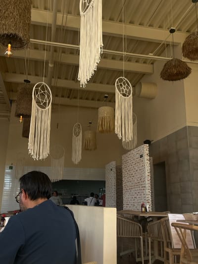 This image captures the interior of a casual dining establishment in Santiago de Querétaro, Mexico, characterized by a bohemian aesthetic. The scene is illuminated by ambient lighting, with a clear view into a service area in the background.

In the foreground, a male subject is seated at a table, facing away from the camera. He possesses short, dark hair and wears eyeglasses. His attire consists of a dark blue long-sleeved shirt, with a dark strap visible over his left shoulder, possibly from a bag. The table he occupies is largely obscured by his posture and the angle.

The establishment's ceiling features exposed white-painted corrugated metal, support beams, and utility pipes. Numerous decorative elements hang from the ceiling at varying lengths. These include multiple large and small macrame dreamcatchers with white string tassels, and several pendant light fixtures. Some light fixtures are covered with woven natural fiber shades, either bell-shaped or cylindrical, in shades of brown or beige, with visible Edison-style bulbs. Other fixtures feature lighter, fringed material.

The walls in the mid-ground and background are painted a light cream color in their upper sections. A white tiled counter or bar runs horizontally across the mid-ground, separating the dining area from a service or kitchen area beyond. Below this line, to the right, are vertically oriented partition walls constructed from white-painted bricks. Further right, the wall features a darker, possibly grey, large-format tile or concrete block pattern. A green exit sign is partially visible on the far left wall.

In the background service area, several indistinct figures are visible. One individual, wearing a light-colored top, stands behind a counter. Other figures are partially obscured, suggesting activity within the service space.

To the right of the foreground subject, several empty tables and chairs are arranged. The chairs are constructed from light-colored woven wood or rattan. On one table, a caddy containing condiment bottles (one red, one brown) is present, along with a white menu resting on a chair. The floor is not distinctly visible but appears to be a light-colored, possibly tiled surface beneath the tables.