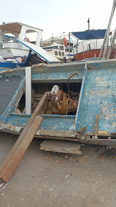 In Mersin, Turkey, the image captures a scene of marine decay in what appears to be a shipyard or boat graveyard. The foreground is dominated by the dilapidated hull of a blue boat, its paint peeling and wood splintered, revealing the rusted remnants of an engine within a gaping aperture. Two wooden planks lie diagonally across the hull's opening, serving as a makeshift bridge or support. Behind this central subject, several other boats are visible, also in various states of disrepair. Some are partially obscured, while others, like the white cabin cruiser in the mid-ground, show signs of abandonment. The sky is overcast, contributing to the somber atmosphere of neglect and decay. The ground is a mixture of dirt and gravel, littered with debris, further emphasizing the disused nature of the location.