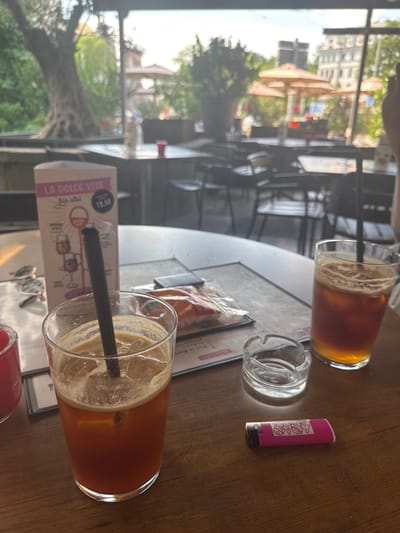 This is a close-up, eye-level shot of a wooden table at an outdoor cafe. In the foreground, there are two glasses of iced coffee, one in focus on the left and another slightly out of focus on the right. An ashtray and a pink lighter are also on the table. In the background, out-of-focus tables and chairs are visible, along with patrons. The cafe appears to be situated in a city, with buildings and trees visible in the distance. The lighting suggests it is either late afternoon or early morning.