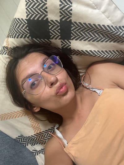 A woman with dark hair and blue eyes, wearing glasses and a peach-colored top, lies on a patterned blanket. Her lips are pursed as she looks at the camera. The blanket has a geometric design of black, white, and tan zigzags. The environment is indoors, with a portion of a blue surface visible at the bottom left. The scene does not provide any visual cues to determine its location within the city of Tryavna, Bulgaria.
