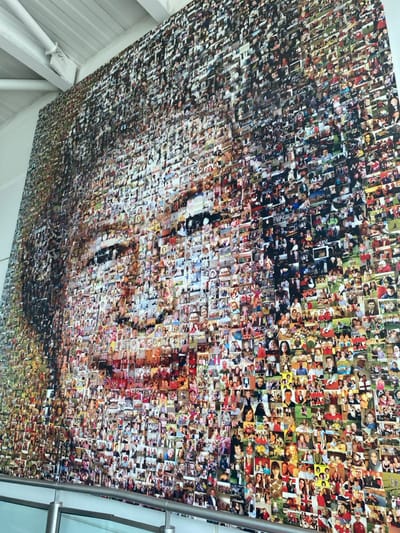 The image displays a large photo mosaic artwork, likely installed on a wall or ceiling in a public indoor setting in Horley, United Kingdom. The mosaic is composed of hundreds, if not thousands, of small, individual photographs. From a distance, these countless smaller images coalesce to form a larger, abstract portrait of a man's face. The man's features appear gentle and somewhat contemplative, with his gaze directed slightly upwards.
Upon closer inspection, each square within the mosaic is a distinct snapshot, predominantly featuring people. These smaller photographs capture a diverse range of subjects, including individuals, couples, families, and children, often engaged in everyday activities, celebrations, or travel. The varied lighting, composition, and photographic styles of the individual pictures suggest they were taken across different periods, creating a vibrant historical collage.
The artwork itself is mounted on a light-colored, possibly white, architectural surface. In the bottom left corner, a white ceiling beam or support structure is visible, while a polished silver metal railing runs along the right side of the frame, suggesting the presence of a staircase, balcony, or elevated walkway in the viewing area. The sheer scale of the mosaic indicates it is a significant art installation within a large building, potentially an airport terminal or a similar public space. The overall impression is one of a community's collective memories and shared human experiences. There is no visible text in the image that can be clearly read.