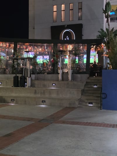 The image captures an outdoor seating area at night, likely a restaurant or bar, situated on the ground floor of a building in Burbank, California, USA. The scene features concrete tiered seating in the foreground, with several embedded lights illuminating the steps. On the second tier of the seating, two patio heaters are prominently positioned.

Behind the seating area, a covered outdoor space is visible, enclosed by transparent material, which appears to be a gathering place for patrons. Inside this space, multiple flat-screen televisions are displaying a soccer match, suggesting a sports-watching venue. The area is decorated with numerous national flags hanging above the seating. Several individuals are seated at tables, some appearing to be engaged in conversation or watching the game. Palm trees and other greenery add to the ambiance. The building facade is visible above the covered area, with a large arched entrance and several windows. To the right of the building, a portion of a blue wall and a railing are visible. The overall impression is a lively urban nighttime scene.