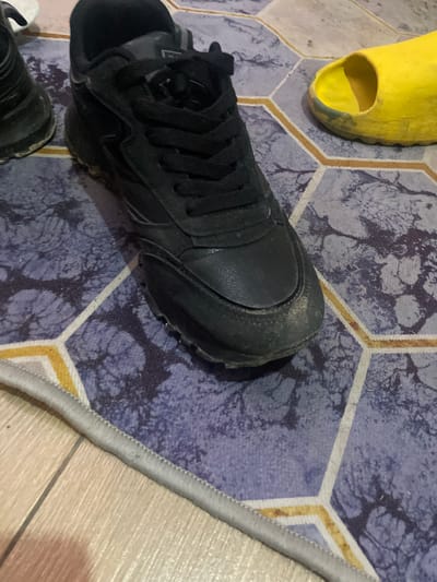 The scene depicts a section of flooring covered by a rug with a purple and grey hexagonal pattern, with a yellow slide and a pair of black sneakers on top of it. The rug's edge is bordered with grey stitching. The flooring beyond the rug is light-colored wood. The location is unknown. No people, actions, or interactions are visible.