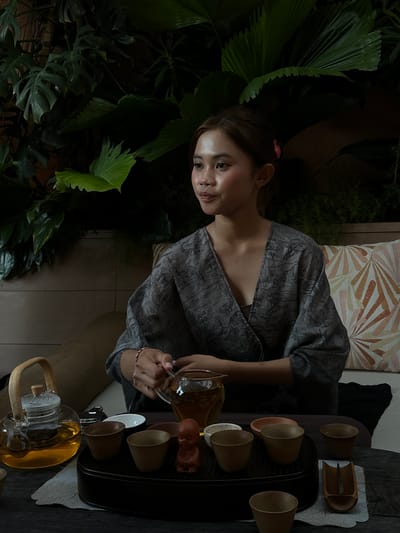 A young woman with long brown hair and fair skin, wearing a grey patterned kimono-style top, pours tea from a glass pitcher into a small cup. She is positioned beside a dark wooden table containing a black tray with several small tea cups and a wooden implement for the tea ceremony. The background includes lush greenery, suggesting a tropical or garden setting. A patterned cushion is visible behind the woman. The scene takes place outdoors, possibly in Pecatu, Indonesia.