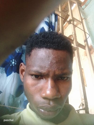 The image shows a close-up selfie of a young Black man with short, dark hair. He is wearing a light-colored shirt. Behind him, there's a window with a curtain featuring a blue floral pattern. To the right of the window, there's a wooden structure, possibly a makeshift support or frame. The lighting suggests an indoor environment with natural light. The image is captioned with the name "paschal." It is not possible to definitively determine if the picture was taken in Nsugpe, Nigeria, based solely on visual information.