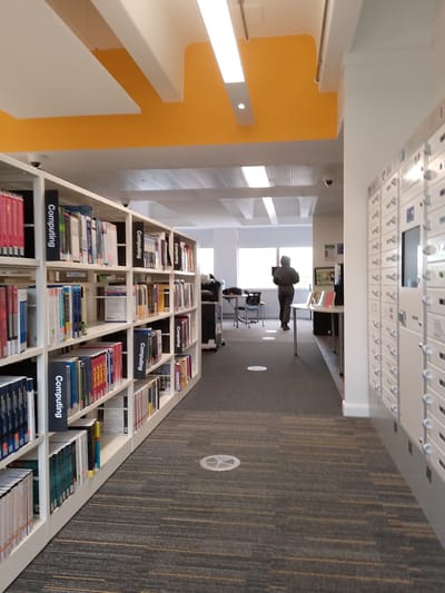 This image depicts the interior of a modern library or study area, likely in Coventry, United Kingdom. The scene is dominated by a long aisle with white, multi-tiered bookshelves on the left, densely packed with books. Many sections on the shelves are clearly marked with black labels reading "Computing," indicating a specialized collection of books. The book spines vary in color, but specific titles are largely illegible.
On the right side of the aisle, there's a wall featuring numerous white locker-style units, arranged in rows. Some units have small screens or interfaces, possibly for self-service or personal item storage. The floor is covered with a patterned grey carpet featuring subtle horizontal stripes of yellow or gold.
Towards the background, a person dressed in dark clothing (hoodie and trousers) stands facing a large window, appearing to interact with a computer screen or device on a table. Additional tables and chairs are visible in the background, along with what appears to be a printer or scanner on a trolley.
The ceiling is a notable feature, with bold yellow panels contrasting with white rectangular structures and integrated strip lighting. Small, dome-shaped security cameras are mounted on the ceiling and above the bookshelves. Natural light entering through the window suggests it is daytime. The overall impression is a well-lit, organized, and quiet environment suitable for study or research.