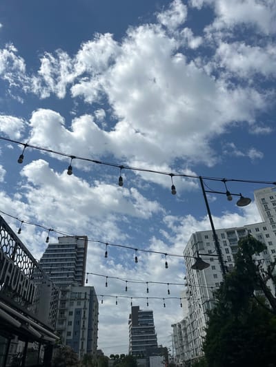 The image shows a view looking up at buildings and a partly cloudy sky in Esenyurt, Turkey. Decorative string lights with bare bulbs are strung between poles. Several tall buildings are visible, including some with visible windows and balconies. A metal structure with a patterned design is partially visible in the foreground. The sky is a mix of blue and white clouds.