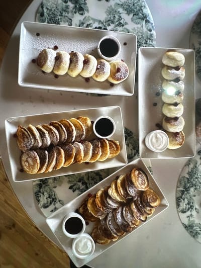 Four white rectangular platters of mini pancakes, dusted with powdered sugar and garnished with a few berries, are arranged on a white table with a floral tablecloth. The platters are positioned diagonally across the frame. The first platter, in the top left, contains five pancakes in a row, with a small bowl of dark sauce to the right. The second platter, below and to the left of the first, holds many more pancakes arranged in rows, with a small bowl of dark sauce to the right and a small bowl of white cream to the lower right. The third platter, to the right of the first, contains six pancakes stacked vertically, with a small bowl of white cream to the bottom right. The fourth platter, in the bottom right, also contains many pancakes arranged in rows, with a small bowl of dark sauce and a small bowl of white cream on the left side. The setting appears to be a breakfast or dessert spread. There are no people or discernible actions depicted in the image. The lighting is soft and diffuse, casting gentle shadows. The overall scene is one of food presentation. The location is not specified in the image.