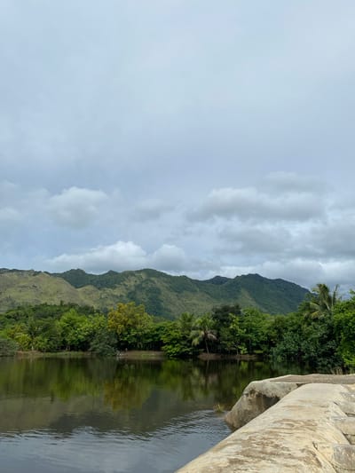 The image shows a tranquil natural scene with a calm body of water in the foreground, reflecting the lush green trees and rolling mountains in the background. The sky is overcast with scattered clouds. In the foreground, large rocks form a pathway or edge along the water. The environment suggests a rural or natural park setting, possibly in Aneuk Paya, Indonesia, characterized by dense vegetation and mountainous terrain. There are no people or animals visible in the image.