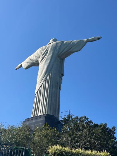 This is a low-angle, full-body shot of the Christ the Redeemer statue in Rio de Janeiro, Brazil, set against a clear, bright blue sky. The statue is made of concrete and soapstone and is depicted with its arms outstretched. The iconic statue is positioned on Corcovado Mountain, overlooking the city. The base of the statue, a dark, rectangular structure, is visible below, partially obscured by lush green foliage and trees. A metal fence and some smaller plants are visible in the foreground. Construction scaffolding can be seen near the base of the statue, suggesting ongoing maintenance or work. The image is captured on a sunny day, with the bright light casting subtle shadows on the statue, highlighting the textures of its robes.