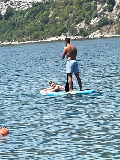 Two individuals are paddleboarding on the calm, blue waters of the Bay of Kotor, with the mountainous, tree-covered shoreline of Risan, Montenegro, in the background.

In the foreground, a person is lying on their back on a light blue inflatable mattress, propped up on their elbows, with their head turned towards the camera and a smile on their face. Their hair is pulled up into a bun, and they are wearing sunglasses and possibly a swimsuit top.

Behind them, a second person stands upright on a blue and yellow paddleboard, holding a paddle. This individual is shirtless, wearing light blue shorts, and facing away from the camera, looking towards the shoreline.

The water is rippled, indicating a gentle breeze. The shoreline features rocky, steep slopes covered in dense green foliage. The overall scene suggests a leisurely day spent on the water in a scenic coastal environment.