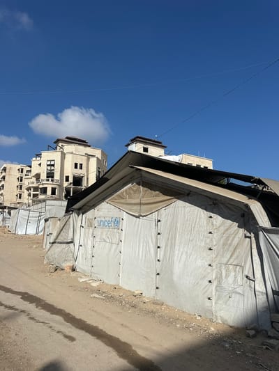 In the city of Gaza, Palestinian Territories, this image depicts a scene of temporary shelters and damaged buildings under a clear blue sky with scattered clouds. In the foreground, a large, white tent with a UNICEF logo prominently displayed on its side is visible. The tent appears to be part of a larger encampment, with other similar white structures stretching into the background. The ground is a mixture of dirt and sand, with some debris scattered around. In the mid-ground and background, several multi-story buildings are seen, some of which appear to be under construction or damaged, with visible signs of disrepair such as broken windows and incomplete facades. Power lines crisscross the sky above the scene. The overall impression is one of a place affected by conflict or displacement, with signs of both immediate aid efforts and the lingering impact of destruction.
