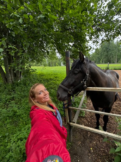 A medium shot captures a woman with blonde hair, wearing a red jacket and a grey t-shirt, smiling and looking at the camera. She is standing next to a dark brown horse, which is behind a wooden fence. The horse is wearing a black halter. The background consists of lush green foliage, trees, and a fenced pasture. The scene appears to be in a rural setting, possibly a farm or stable. The lighting suggests an overcast day. The image was taken in the city of Mars, Russia.