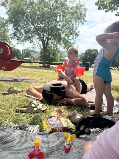 A family is enjoying a sunny day at a park, possibly near a swimming area given the presence of children in swimwear and arm floaties. A man lies on his back on a picnic blanket, with a young girl sitting on his chest, both appearing happy and engaged. Another child, a girl wearing a blue swimsuit, stands nearby, also looking happy. The scene is set outdoors on a grassy area with trees in the background. Several objects are scattered on the blanket and grass, including sandals, a bag of snacks, and children's toys. The weather appears to be warm and sunny, with a blue sky and scattered clouds visible. The overall atmosphere is relaxed and joyful, suggesting a pleasant family outing. There is no visible text in the image.