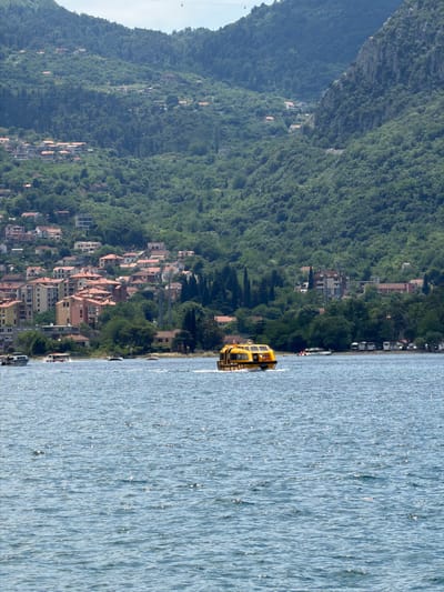 A high-angle shot depicts a body of water with a small yellow boat near the shore of Dobrota, Montenegro. The shoreline is densely populated with buildings of various styles and sizes, interspersed with greenery. A tall, tree-covered mountain looms in the background. The sky is partially visible in the upper left corner.