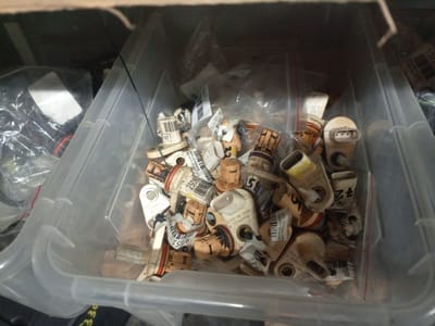 The image shows a clear plastic storage bin filled with numerous small, beige and brown electronic components, possibly automotive sensors. Many of these components have small white labels with black text and barcodes attached to them. The bin is situated on a dark, flat surface, and the background is out of focus, suggesting an indoor setting, possibly a garage or storage area. The lighting is dim. The overall impression is one of organized clutter, with the components piled loosely within the bin. No specific location in Oktyabr'skii, Russia is discernible from the image itself.