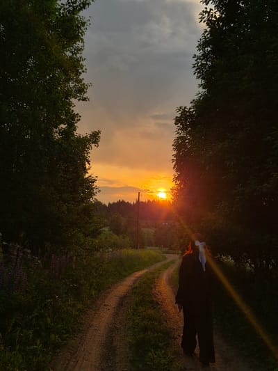 An individual, viewed from behind, walks on a dirt path through a rural area at sunset. The path curves to the left, bordered by lush green vegetation and trees on either side. The setting sun casts a warm, orange glow across the sky, illuminating the clouds and creating a lens flare effect visible in the lower right portion of the image. In the distance, a line of trees forms a dark silhouette against the vibrant sky. The person is wearing dark clothing and has a white ribbon tied in their hair. The environment appears to be a natural landscape, possibly on the outskirts of a village, given the presence of what might be scattered structures or utility poles in the far distance. The specific location in Дойная, Russia cannot be determined from the visual information alone.