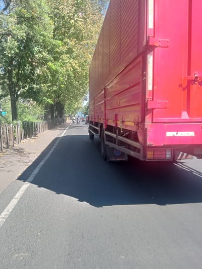 A red truck is shown from a low angle, driving on a road in Waru, Indonesia. The truck is in the right half of the frame, its large, red cargo container dominating the view. The road is asphalt, with a white line marking the lane on the left side. A sidewalk with a wooden fence is visible to the left of the road, lined with trees that cast long shadows. Further down the road, there are a few motorcycles and cars. The sky is clear and blue, suggesting a sunny day. The image appears to have been taken from a moving vehicle, as indicated by the motion blur and low perspective.