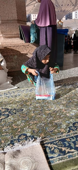 The image depicts an outdoor area within the courtyard of the Masjid an-Nabawi (Prophet's Mosque) in Medina, Saudi Arabia. The ground is covered by a patterned green and blue carpet with floral motifs, with a corner of a beige prayer rug with white fringe visible in the lower left foreground. A child, identifiable as female, wearing a black hijab and a dark long-sleeved top with green and orange patterned cuffs, is squatting on the carpet. She is holding open a transparent blue plastic bag with both hands, looking down at items contained within, which appear to have red and white packaging. Behind and to the right of the child, an adult in a long purple abaya and matching head covering stands, handling a teal-colored bag near a blue waste receptacle or storage bin. To the far left, a person wearing a white garment is partially visible, seated on the carpet. In the background, large white retractable umbrellas, distinctive to the mosque's architecture, are open above, with modern buildings visible beyond them. The scene is brightly lit by natural sunlight, casting distinct shadows.