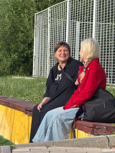 Two women are sitting on a bench in Shcherbinka, Russia. The woman on the left is wearing a black dress and has short brown hair. She is smiling and looking to her right. The woman on the right is wearing a red jacket and has blonde hair in a braid. She is looking at the woman on the left. Behind them is a white metal fence and green trees. The bench is made of wood and is painted red on the top and yellow on the side. The ground in front of the bench is made of concrete and gravel.