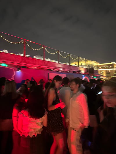 A crowded outdoor rooftop bar or club at night in Shanghai, China, is depicted. A group of young adults are socializing and dancing under red and pink lighting. In the background, illuminated buildings and strings of yellow lights suggest an urban setting. The sky is dark with clouds. The image is blurry due to low light conditions and movement.