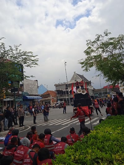 The image shows a street scene in Yogyakarta, Indonesia (latitude: -7.783, longitude: 110.367). A crowd of people, mostly wearing red shirts and vests, are gathered in front of a white building with a red banner and a sound system on top of a truck. There are several individuals standing on top of the truck, including one who is addressing the crowd through a microphone. Other individuals stand near the truck, while the majority of the crowd sits on the ground facing the truck. The street is crossed with a zebra crossing, and there are several vehicles parked on the side of the street. There are trees and foliage on either side of the street. The sky is partly cloudy. The scene appears to be a public gathering of people with red shirts and vests. They are likely attending a rally or political event.