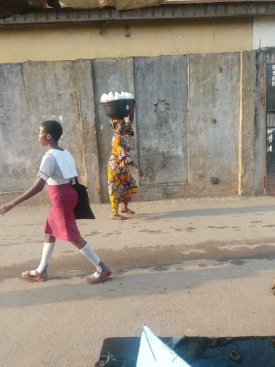 A woman wearing a yellow and brown patterned outfit walks along a sidewalk carrying a large black bowl filled with white items on her head. In the foreground, a young woman in a school uniform with a white top, maroon skirt, white socks, and brown sandals walks in the opposite direction. They both walk alongside a weathered gray wall with a few decorative star patterns. The ground is gray and slightly uneven.
