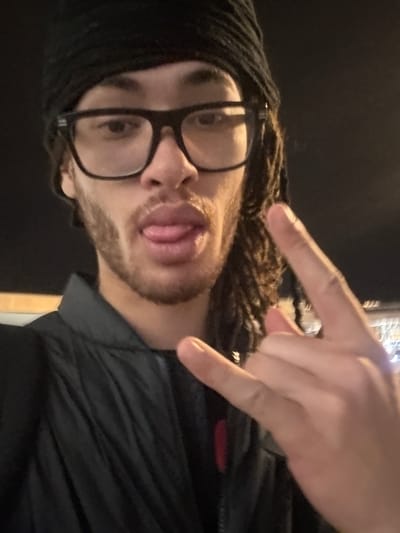 A man is posing for a selfie-style photograph. He is wearing glasses and a black beanie, and has dreadlocks. He is sticking his tongue out and making a hand gesture. He is wearing a dark jacket. The background is dark. The location of the image within Paray-Vieille-Poste, France is indeterminate.