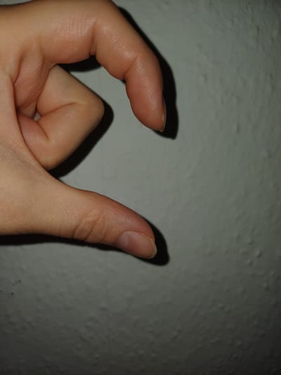 This media file contains a close-up shot of a person's hand against a textured, light-colored wall. The hand is positioned in the lower left quadrant of the image. The thumb is extended upwards, while the index finger is bent, creating a C-shape with the other fingers. The lighting suggests a shadow is cast by the hand onto the wall, indicating the light source is likely to the left and slightly above the hand. The texture of the wall appears to be rough or bumpy. The overall tone of the image is neutral, with no explicit information about the time of day, weather, or specific location beyond the given context. No text is visible in the image. The focus is solely on the hand and its interaction with the background.
