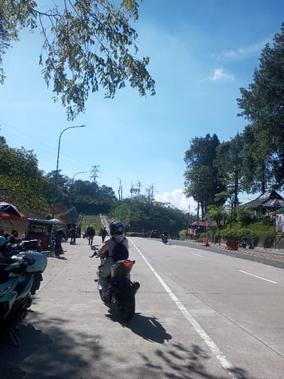 The image depicts a sunlit roadside scene in a hilly, vegetated area identified as Ka Thaung (Upper), Myanmar. In the mid-ground, a person wearing a full-face helmet, light-colored jacket, and dark backpack is positioned on a black motorcycle with a visible red tail light, facing away from the camera, on the paved shoulder of a two-lane asphalt road. Overhead, a leafy tree branch casts shadows onto the concrete. To the immediate left of the central motorcycle, a light-turquoise scooter and other motorcycles are parked. Further left, a series of open-air vendor stalls with dark umbrellas and awnings line the roadside, frequented by multiple individuals. Beyond the stalls and ascending a steep, tree-covered hillside, a concrete staircase or ramp is visible, traversed by several people. Electrical power lines and transmission towers are present atop the distant hills. The road extends into the distance on the right, marked with a solid white line for the shoulder and a dashed white center line. The right side of the road features a patterned sidewalk with orange traffic barriers and tall evergreen trees. A building with a dark, multi-tiered roof is partially visible behind the trees on the far right. The sky is bright blue with scattered white clouds. Distant motorcycles are also observable on the road.