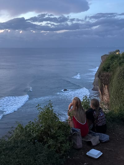 Two individuals are sitting on a cliff edge overlooking the ocean. The cliff is covered with vegetation. One person is wearing a black shirt with words printed on it, and the other is wearing a red top with white straps. A white styrofoam container, a gray bag, a purse, and a piece of clothing are near them. The ocean is visible below the cliff, with waves crashing against the rocks. The sky is cloudy. Based on the setting, this scene is likely in Uluwatu, Indonesia.