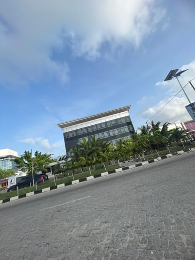 The image depicts a street scene in Ilado, Nigeria, angled from the ground upwards towards a cloudy sky. The road is paved with cobblestones and has a black-and-white painted curb. A modern office building is visible, surrounded by palm trees and other greenery. A traffic sign and a parked car are on the side of the road.