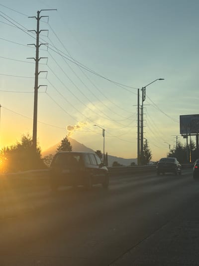 An eye-level shot captures a highway at sunset. Power lines stretch across a blue sky, while silhouettes of distant mountains and trees sit at the golden horizon. Two cars are visible on the road. The overall image is partially obscured by a bright, golden sun flare.