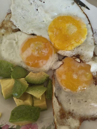A close-up, overhead shot displays a plate of food containing four fried eggs and diced avocado. Three of the eggs are whole with visible yolks, while the fourth is partially obscured. The avocado is cut into irregular cubes and scattered around the eggs. The plate has a floral pattern on its rim, with pink flowers visible. The food appears to be a breakfast meal. There is no information to suggest the location is in Pletena, Bulgaria.